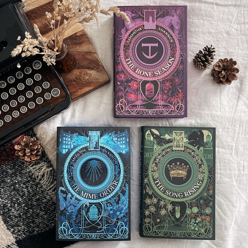 Three decorative book-style boxes or boxes with illustrated covers arranged on a white fabric surface, alongside a vintage typewriter, dried flowers, and pinecones. The top box reads 'The Bone Season' and is purple with pink detailing; the bottom left reads 'The Mime Order' and is blue with teal detailing; the bottom right reads 'The Song Rising' and is green with gold detailing. image
