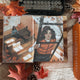 An open illustrated notebook or journal laid on a fabric surface with autumn leaves, pinecones, and a typewriter around it. The artwork shows two women in an indoor scene with warm orange, brown, black, and white tones.