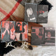 A set of illustrated art cards or prints arranged on a wooden block over a plaid blanket and white fabric. The artwork uses a limited palette of black, gray, red, and beige, depicting stylized portraits and scenes.